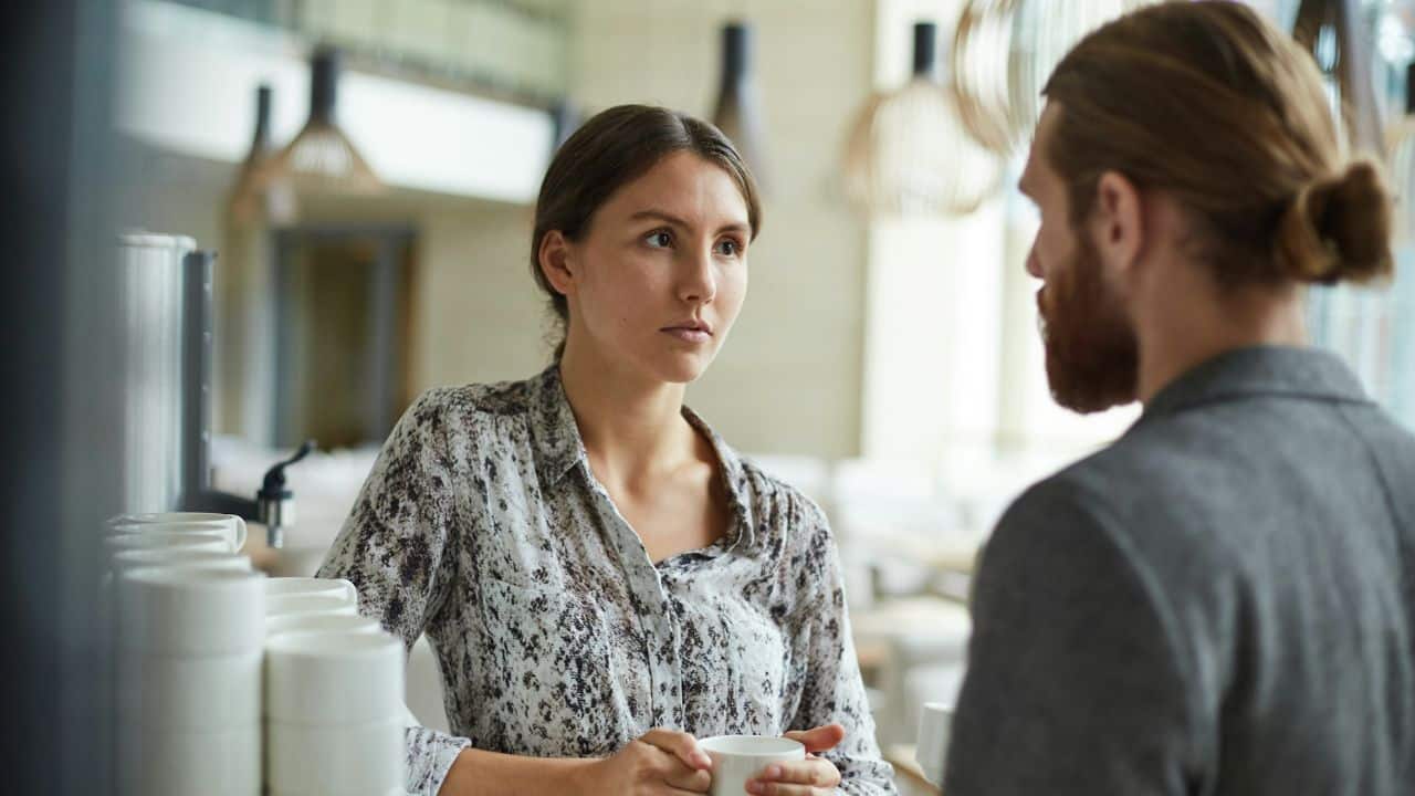 A woman in a patterned shirt holds a cup and looks directly at a bearded man.