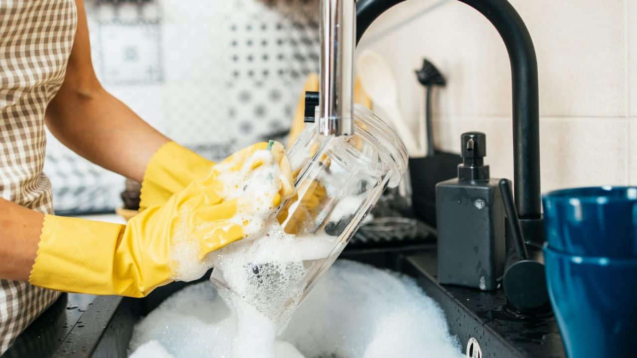 Hands in yellow rubber gloves wash a glass jar in a sink filled with soapy water.