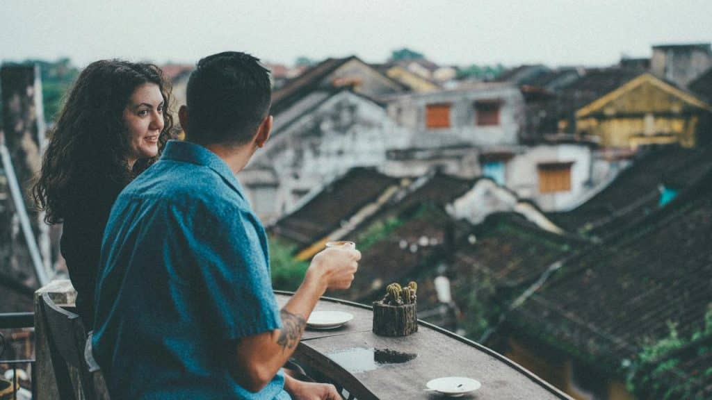 A couple chatting over drinks on a balcony overlooking old rooftops.