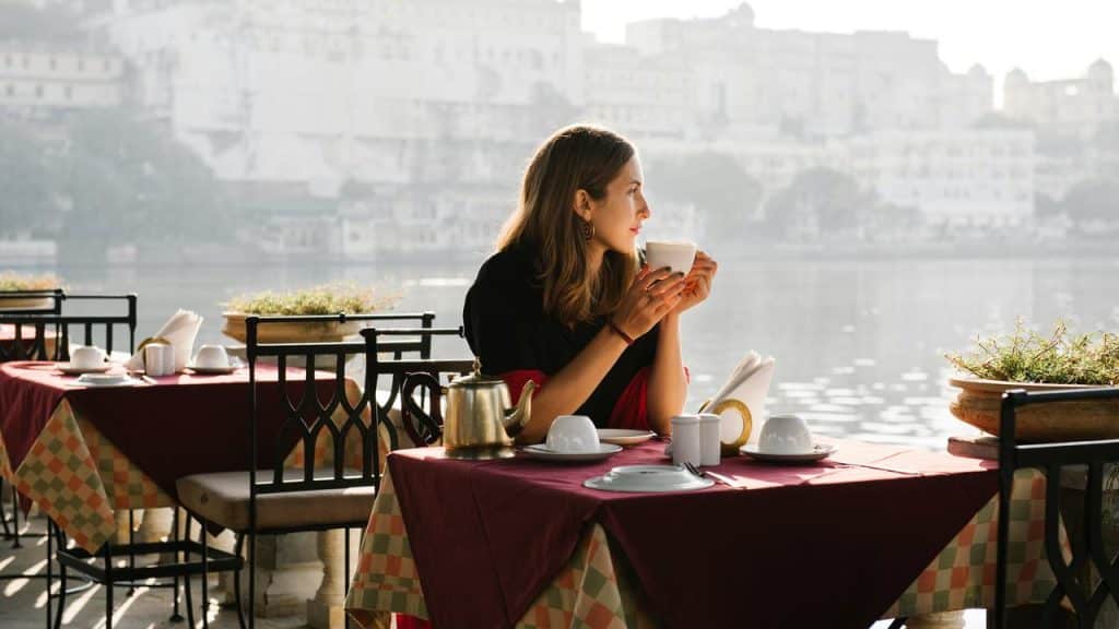 A woman sitting at an outdoor café by the water, holding a cup and gazing ahead.
