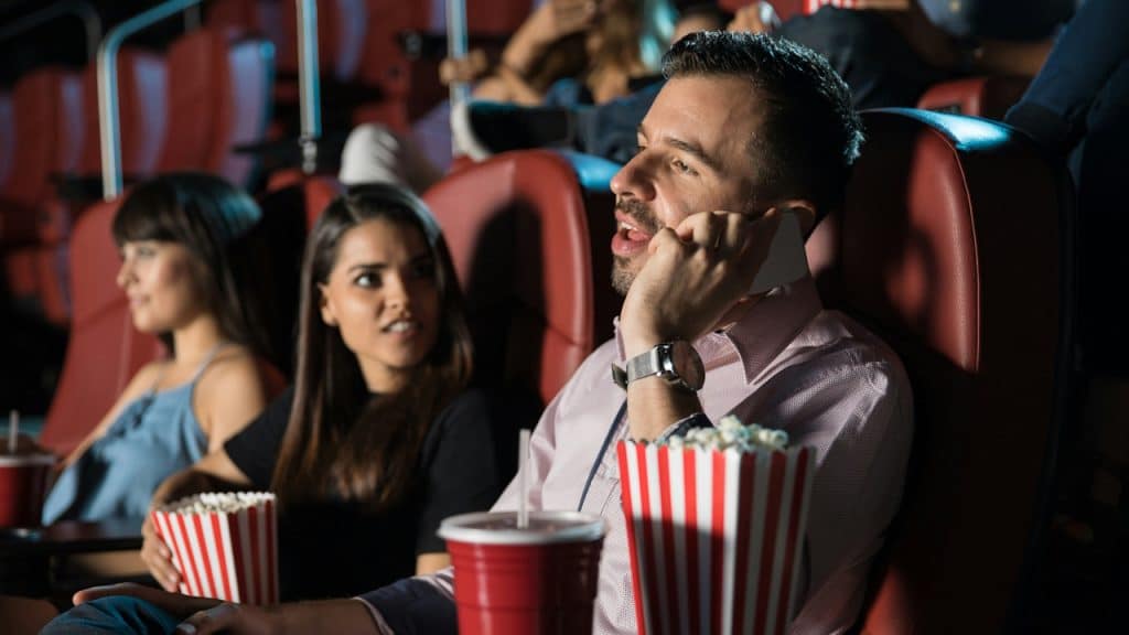 A man talking on his phone while he and his girlfriend are watching a movie in a cinema.