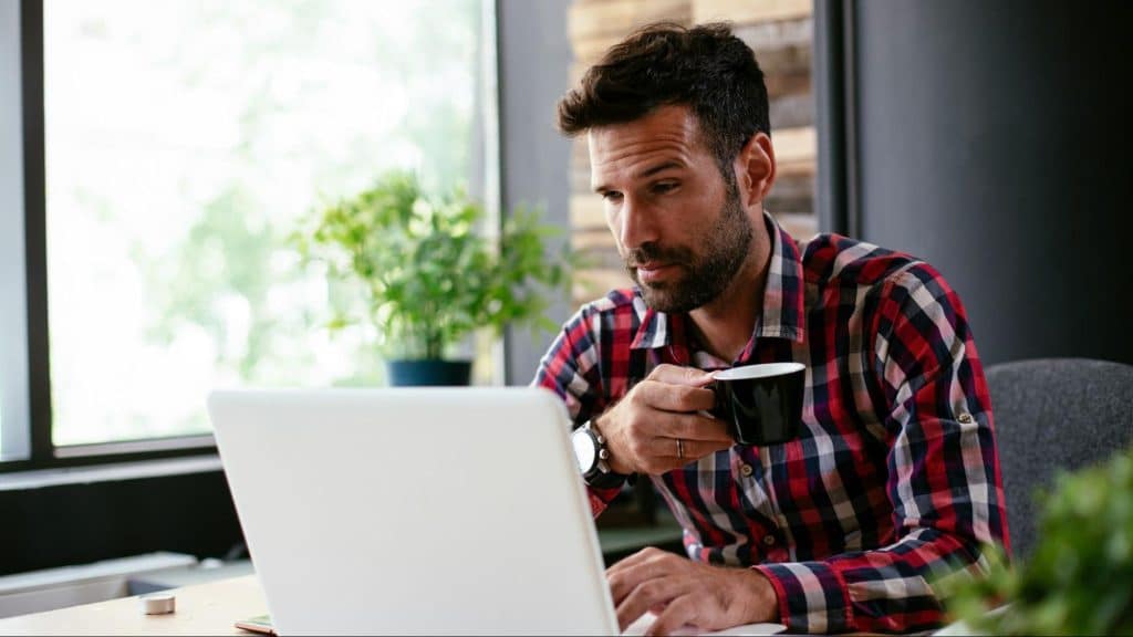 A man looking at the laptop screen while drinking coffee