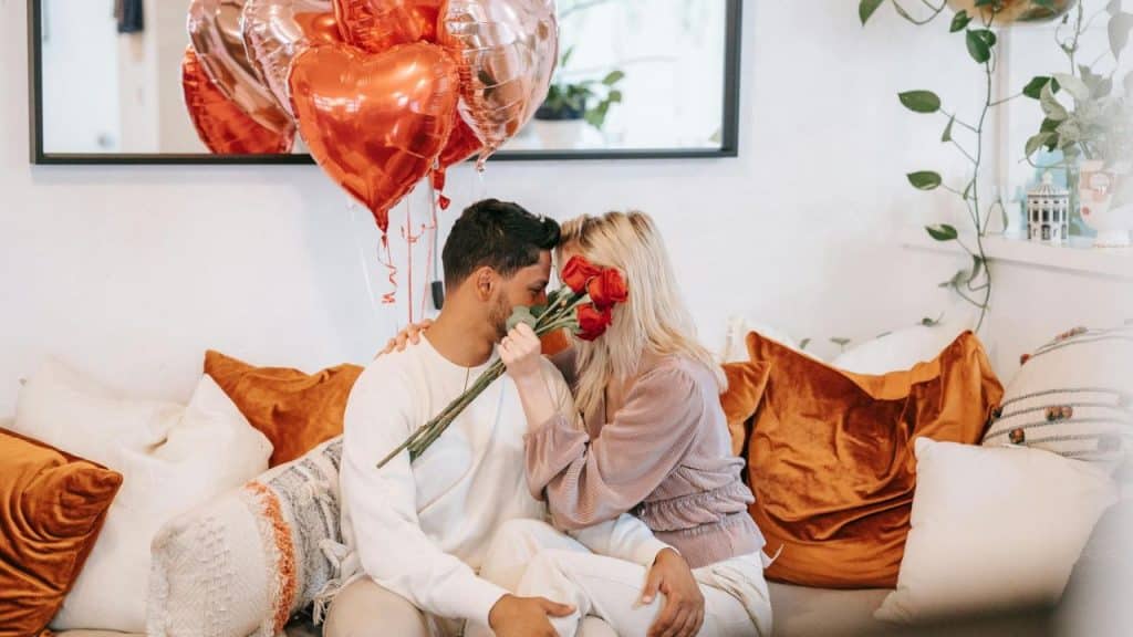 A couple sits on a sofa behind a bouquet of roses near heart-shaped balloons.
