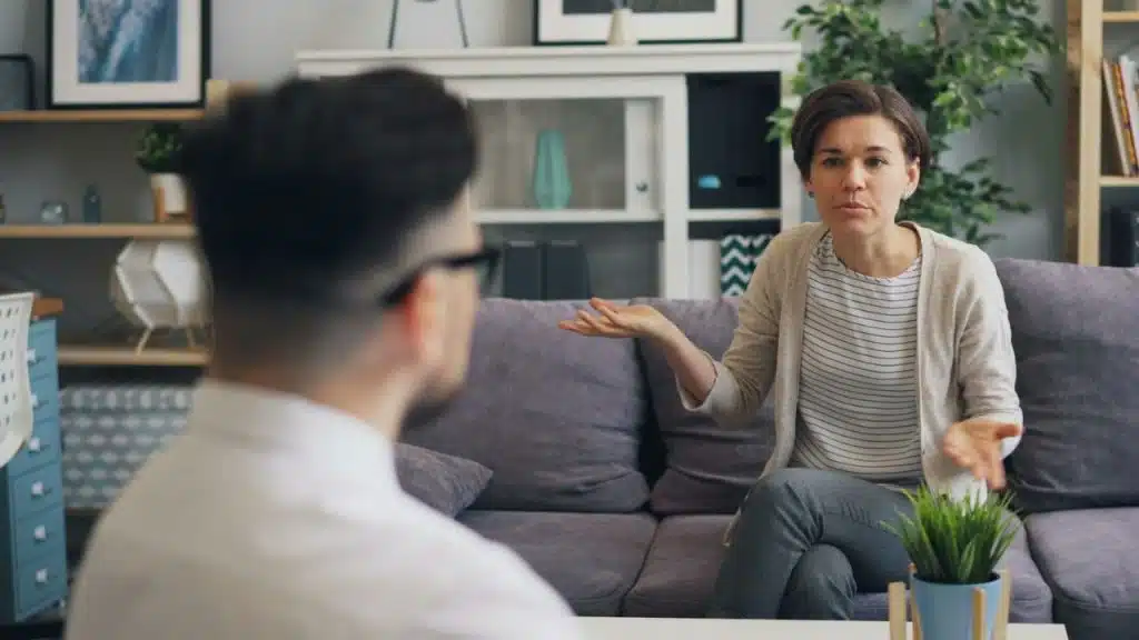 A woman sits on a couch with her hands open while speaking to a man.