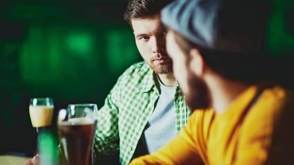 Two men sit at a bar with glasses of beer, one looking directly at the other.