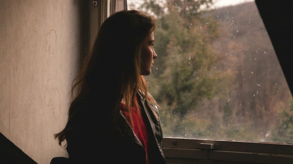 A woman in a black jacket looks out a rainy window toward a forest