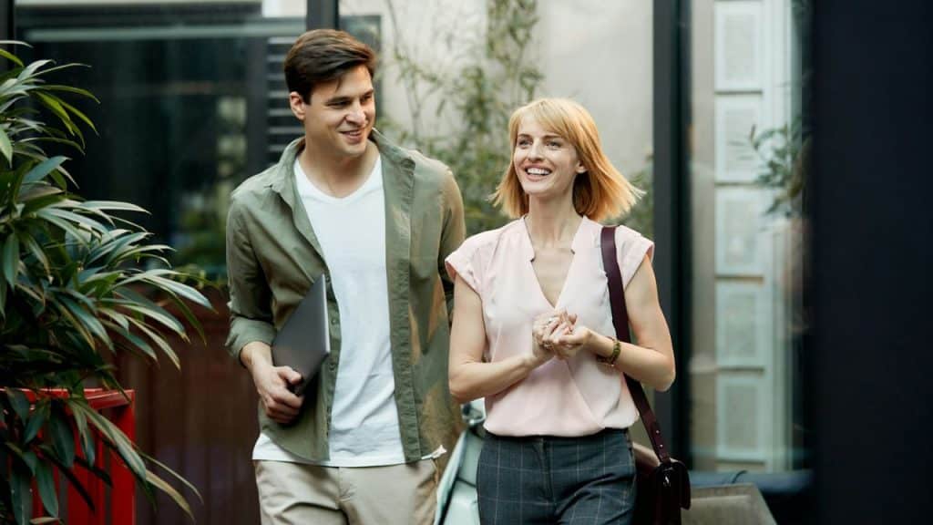 A woman and a man carrying a laptop walk together and smile in an outdoor setting.