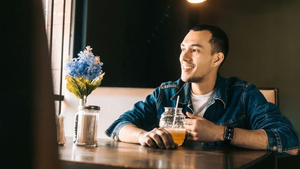 A man in a denim jacket sits at a table, smiling while holding a jar drink.