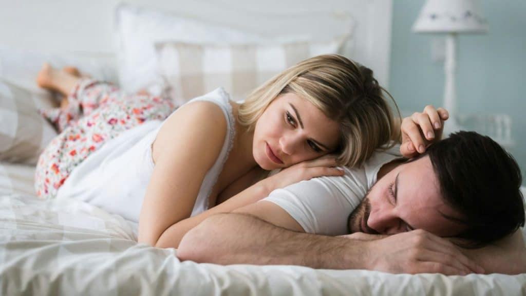 A woman lies on a bed, resting her head on a sleeping man’s back while looking away.