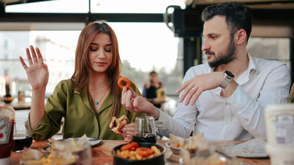 A man and woman sit at a table with food while he holds an onion ring.