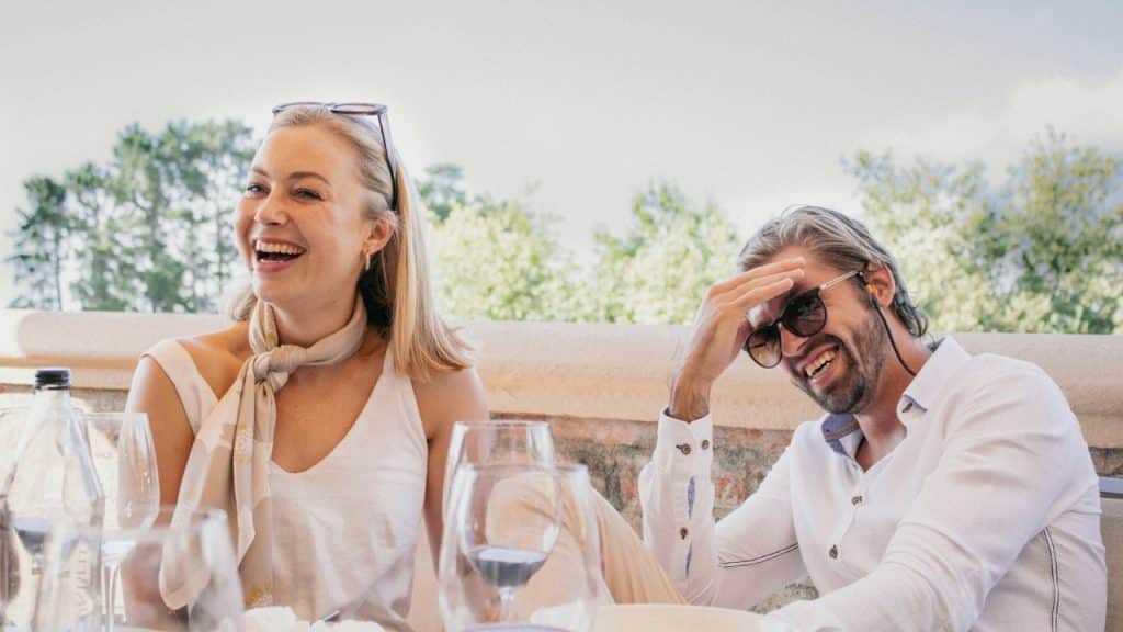 A man and woman sit at an outdoor table laughing together while drinking wine.