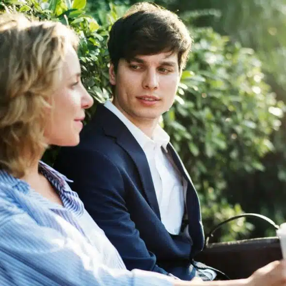 A man and woman sit on a park bench talking while he holds a cup.