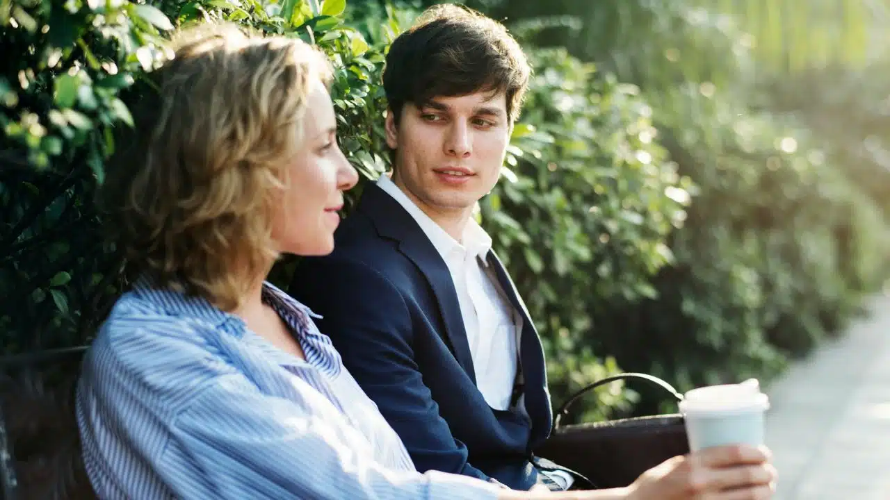 A man and woman sit on a park bench talking while he holds a cup.