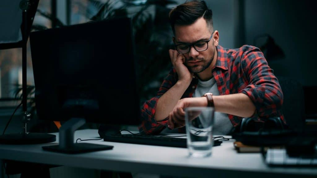 A man in glasses at a desk looking at his watch beside a computer.