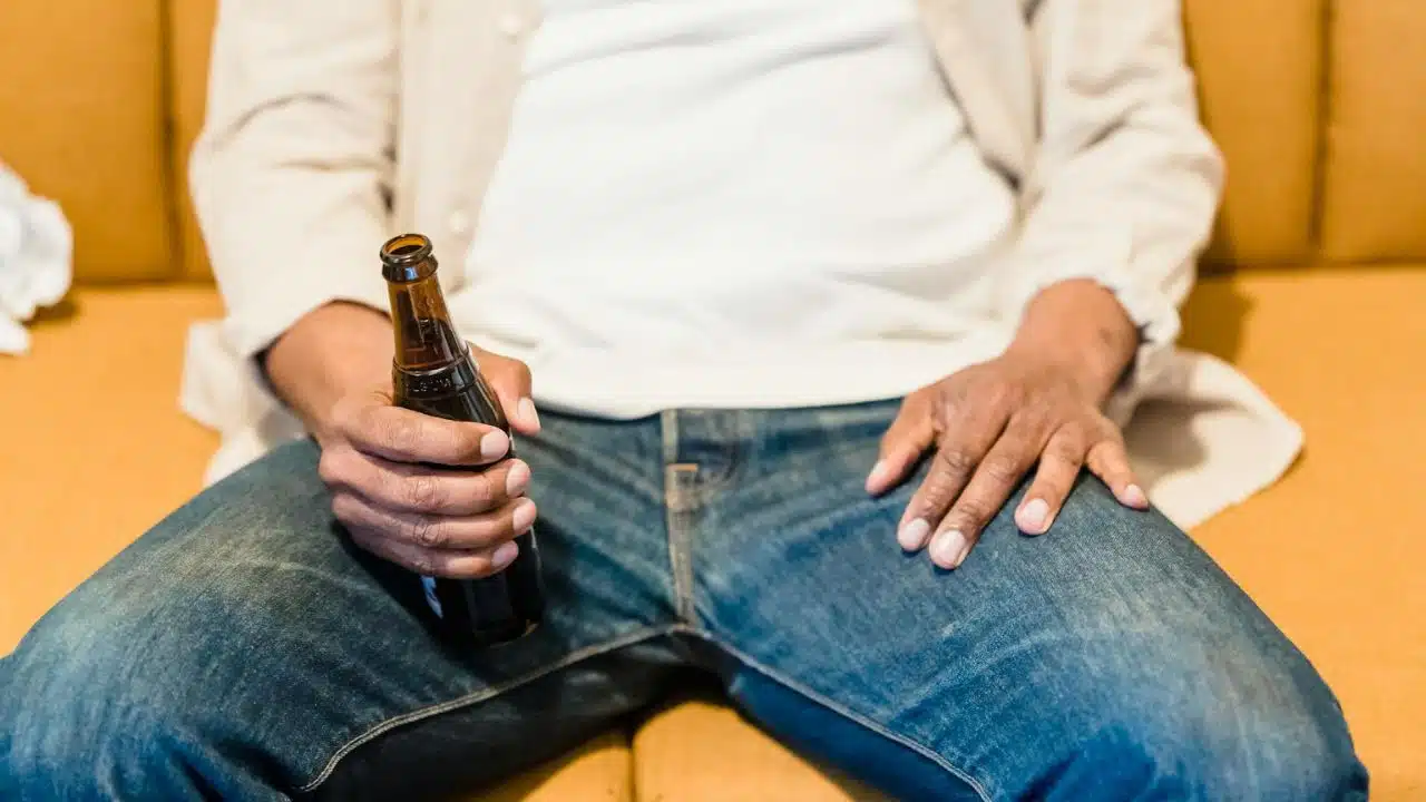 A person in jeans sits on a sofa holding a brown glass bottle in their hand.