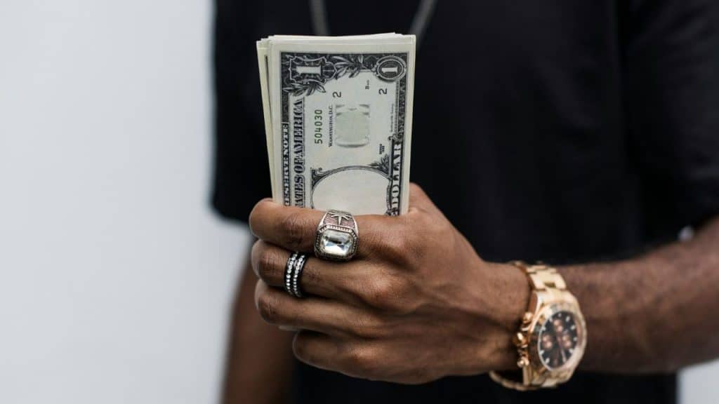A person with rings and a gold watch holds a thick stack of folded one-dollar bills.