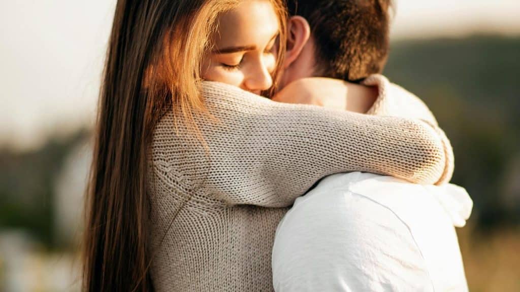 A woman hugging a man closely with her eyes closed in warm outdoor light.