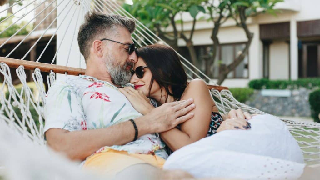 A bearded man and a woman cuddle together in a white rope hammock outdoors.