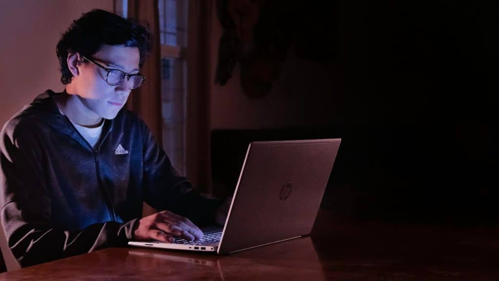 A person wearing glasses and a hoodie uses a laptop in a dark room.