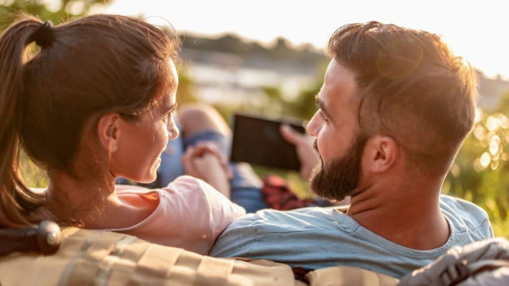 A woman with a ponytail and a bearded man sit closely together outdoors at sunset.
