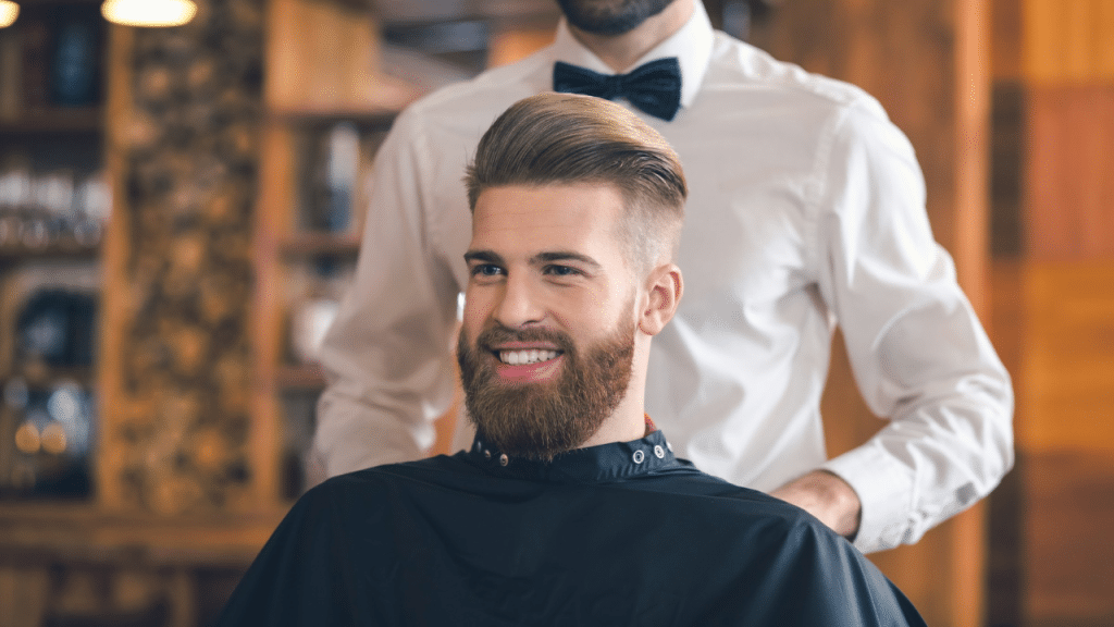 A bearded man with styled hair smiles while sitting in a barber chair wearing a cape.