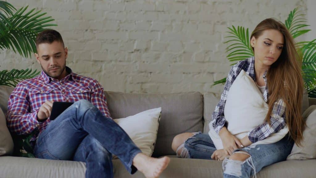 A man looks at a tablet while a woman sits beside him holding a pillow.