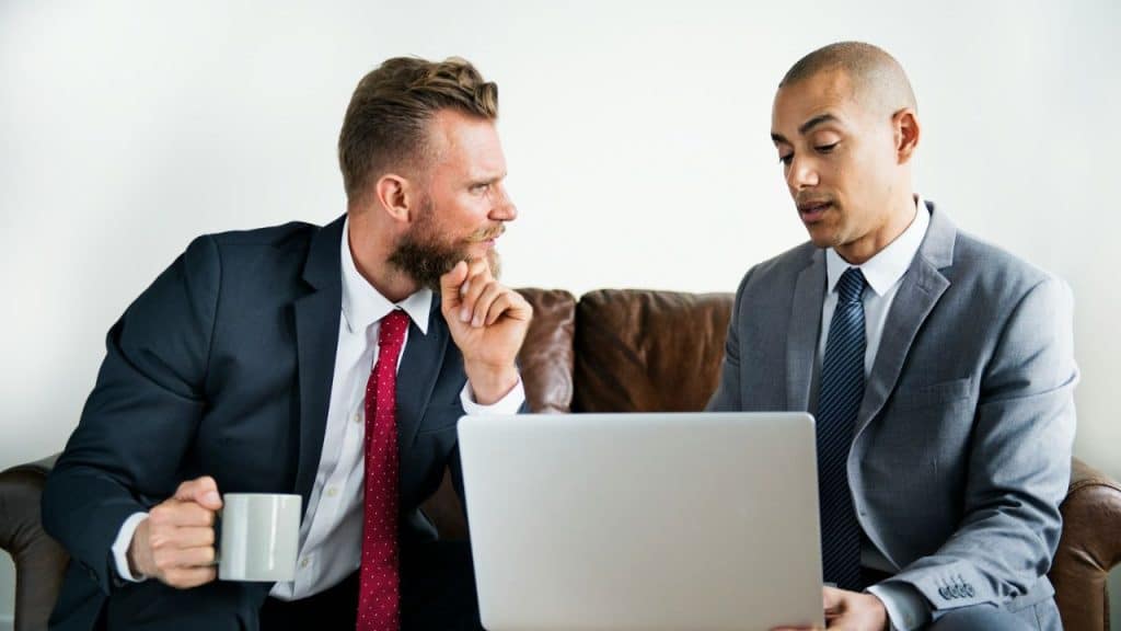 Two men in business suits sit on a leather couch looking at a laptop computer.