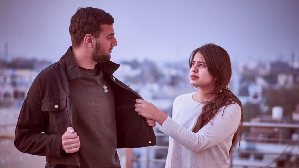A woman pulls on the jacket of a man who is looking back at her outdoors.