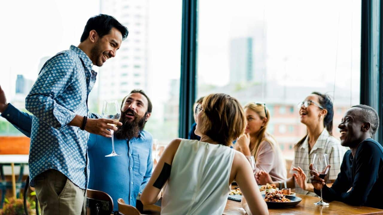 A group of diverse friends laughs and talks together around a dinner table with drinks.