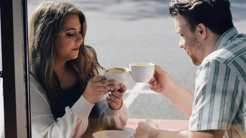 A man and woman sit at a table, tapping their white coffee mugs together.
