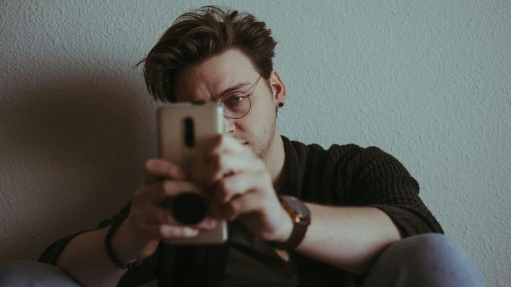 A man with glasses sits against a wall and looks at his mobile phone.
