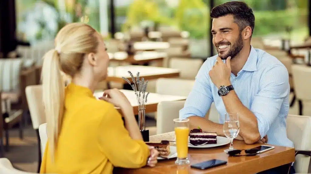 A smiling man and blonde woman sit at a cafe table with cake and drinks.