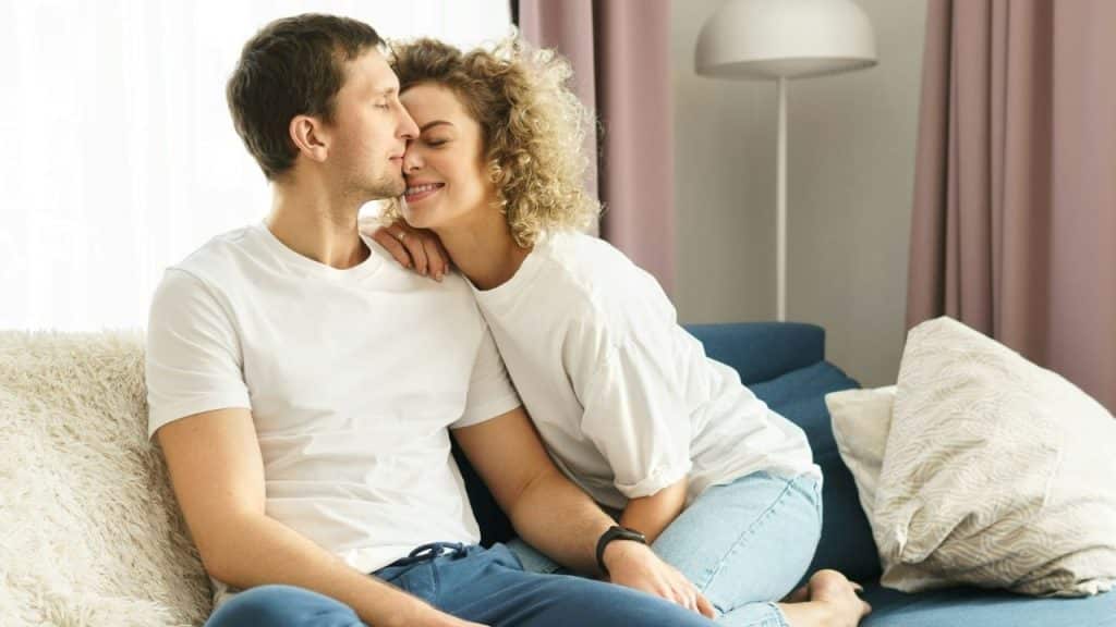 A woman with curly hair leans her head against a man sitting on a sofa.