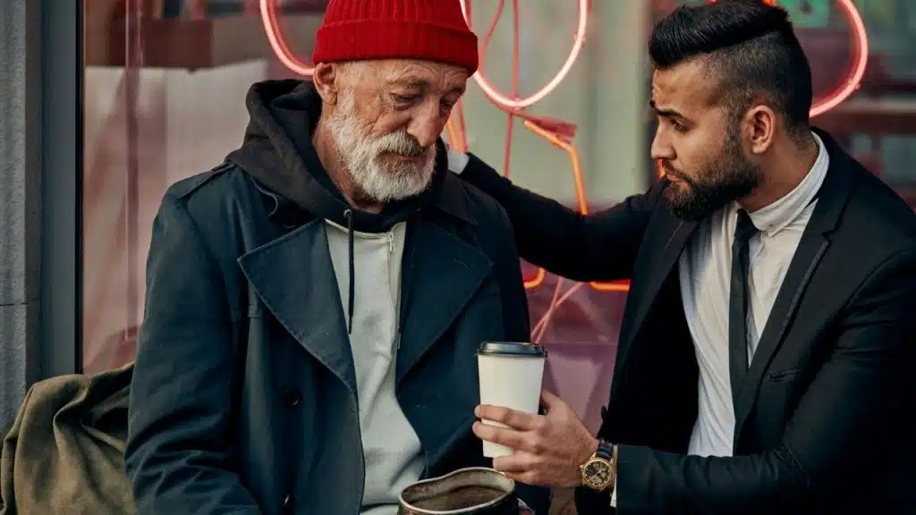 A man in a suit hands a cup of coffee to an older man in a red beanie.