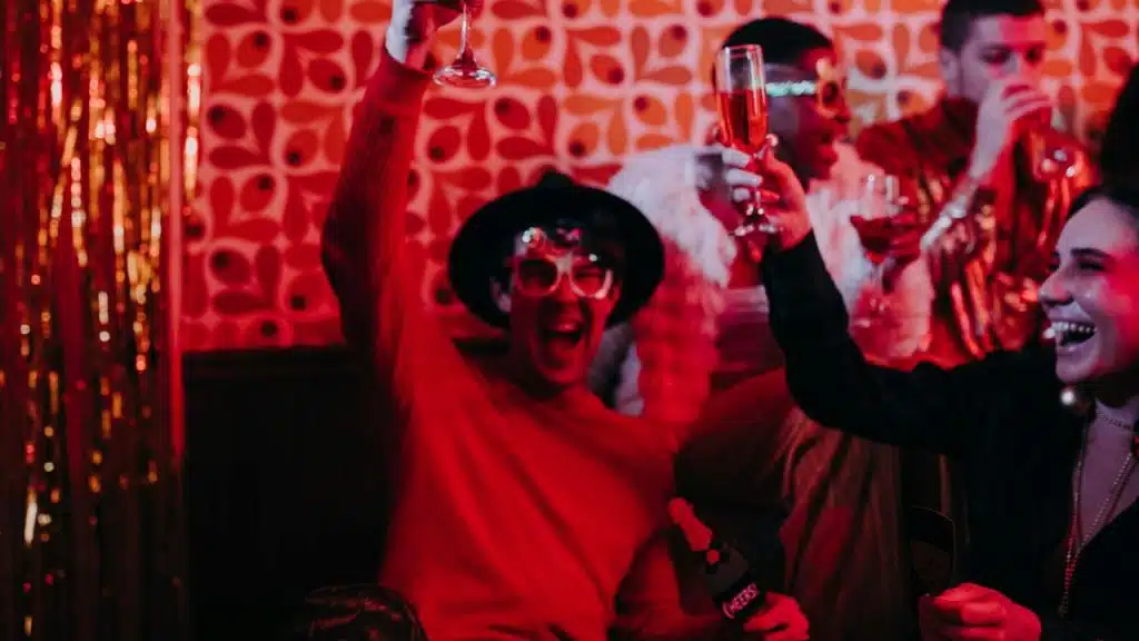 People in festive clothing and glasses cheer and hold up drinks in a red-lit room.