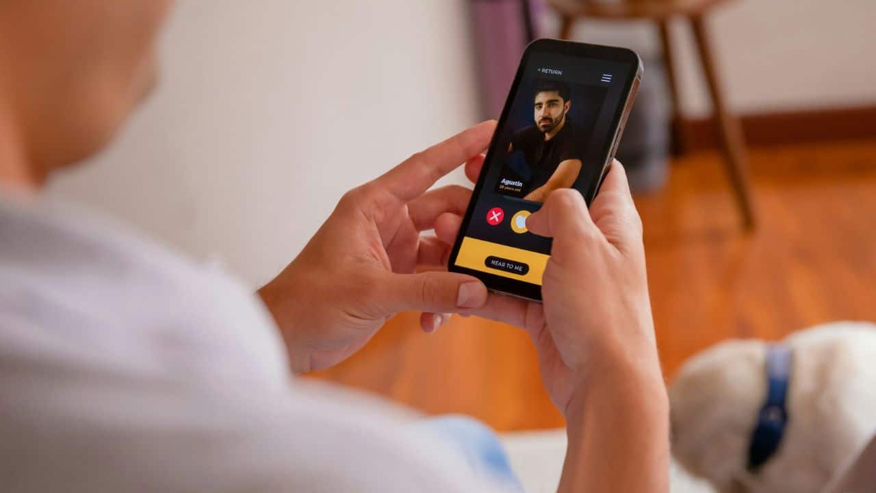 A person's hands hold a smartphone displaying a dating profile with a yellow interface.