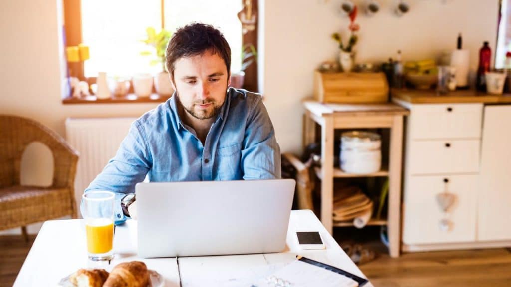 A man sits at a table with a croissant and orange juice while using a laptop.