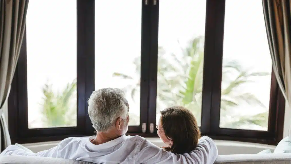 An older couple sits on a couch, looking out a window at palm trees.