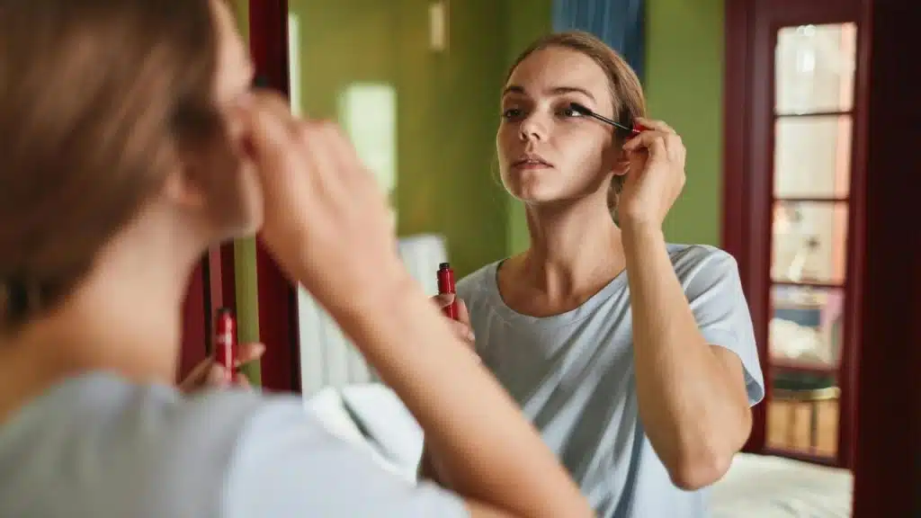 A woman applying mascara while looking into a mirror.