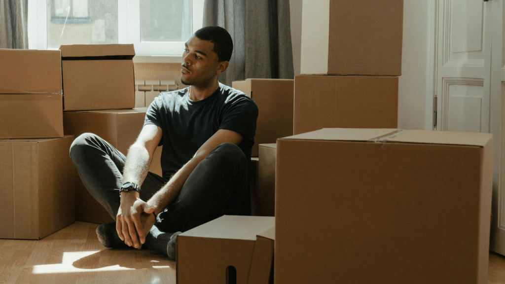 A man sits cross-legged on a wood floor surrounded by several stacked cardboard moving boxes.