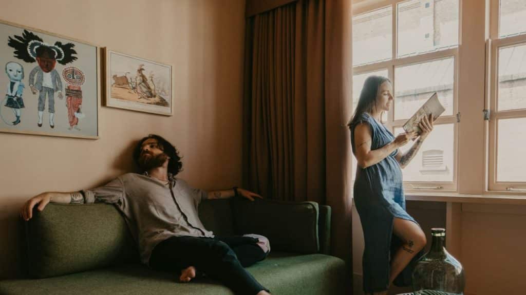 A man reclines on a green sofa while a woman stands by a window reading.