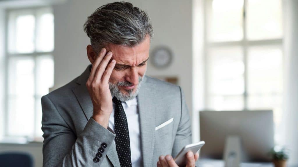A middle-aged man in a suit looks at his phone with a distressed facial expression.