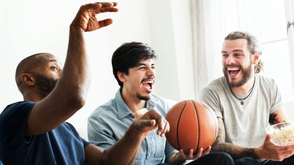 Three men laugh together while one holds a basketball and another holds a bowl of popcorn.