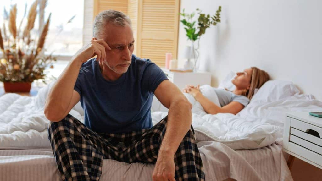 A man in plaid pants sits on a bed looking away from a reclining woman.