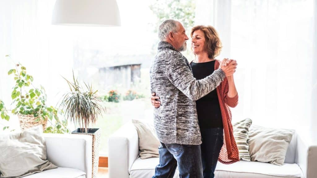 An older man and woman hold hands and dance together in a bright living room.