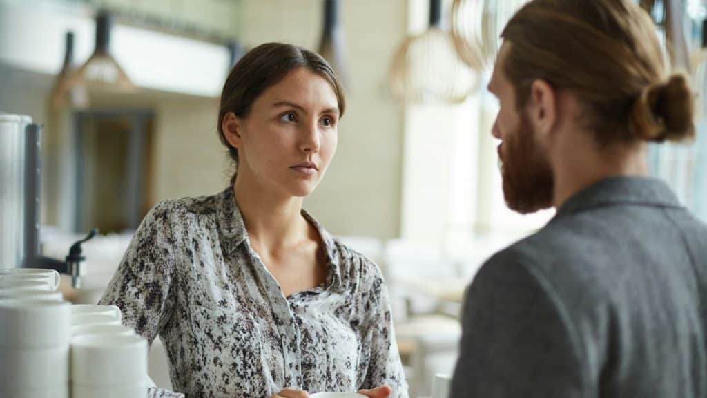 A woman in a patterned blouse looks toward a man with his hair in a bun.
