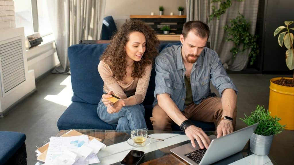 A man and woman sit on a blue sofa while looking at a laptop and credit card.