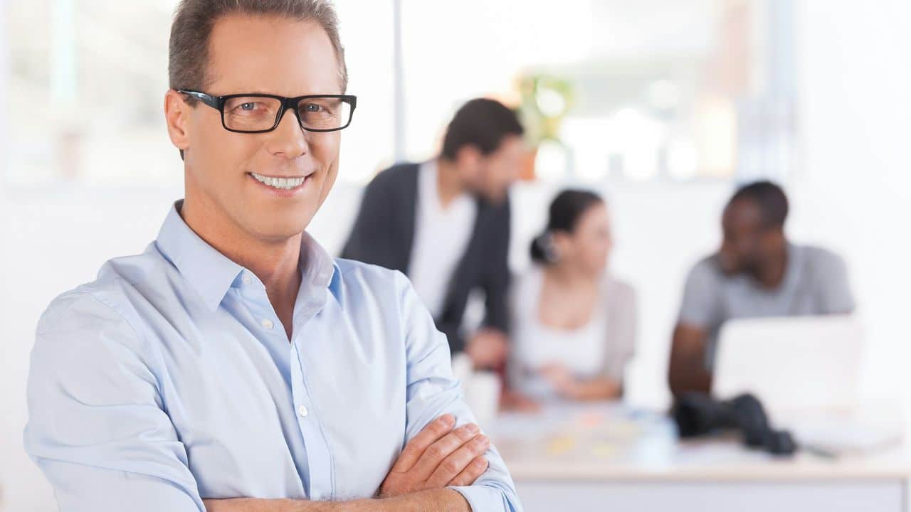 A smiling man in a light blue shirt stands with arms crossed in an office.
