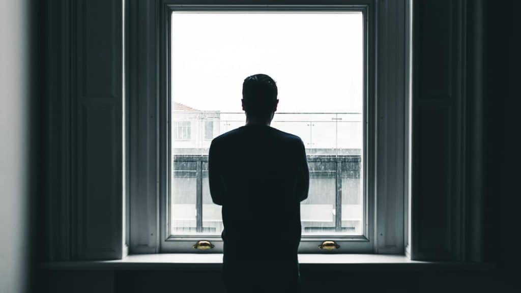 A silhouetted person standing indoors and looking out through a large window.