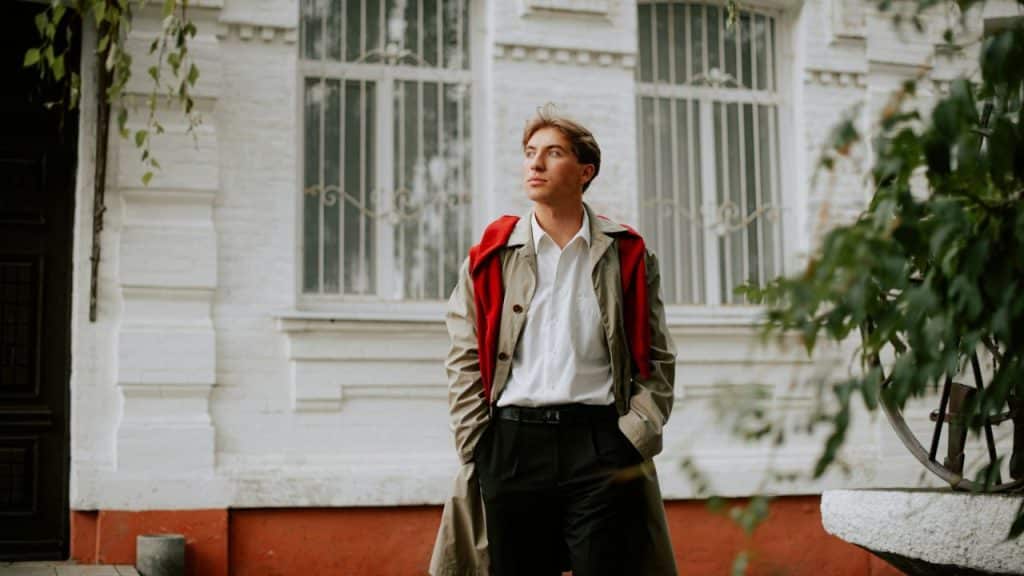 A man in a trench coat and red sweater walks past a white building.
